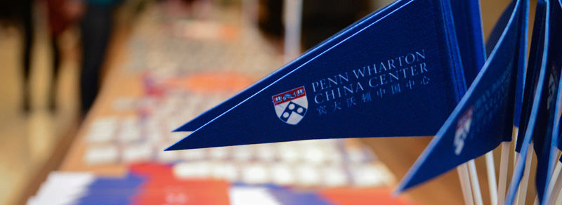 A display of blue pennants featuring the Penn Wharton China Center logo and text, with a blurred background of tables.