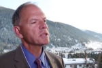 A person in a suit and tie is speaking with a snowy mountain landscape and a town visible in the background.